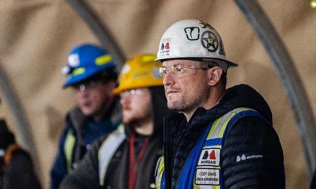 A close-up photo of a male construction worker in a white hard hat sitting next to two other construction workers in a brown shelter.
