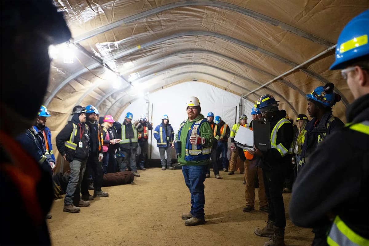 A large group of construction workers standing in a circle under a brown shelter.