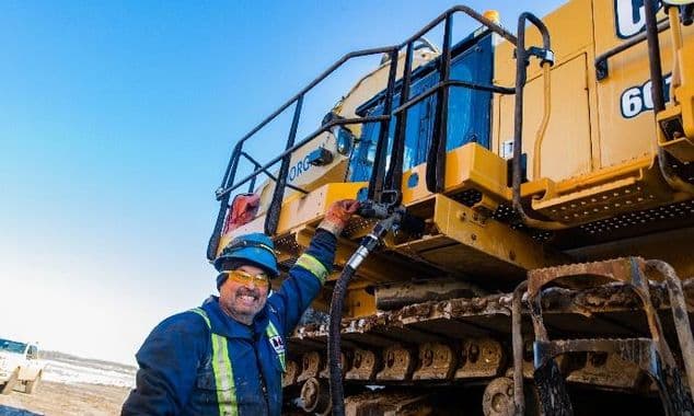 A bearded male construction worker in blue reflective coveralls and a blue hard hat standing next to yellow machinery.
