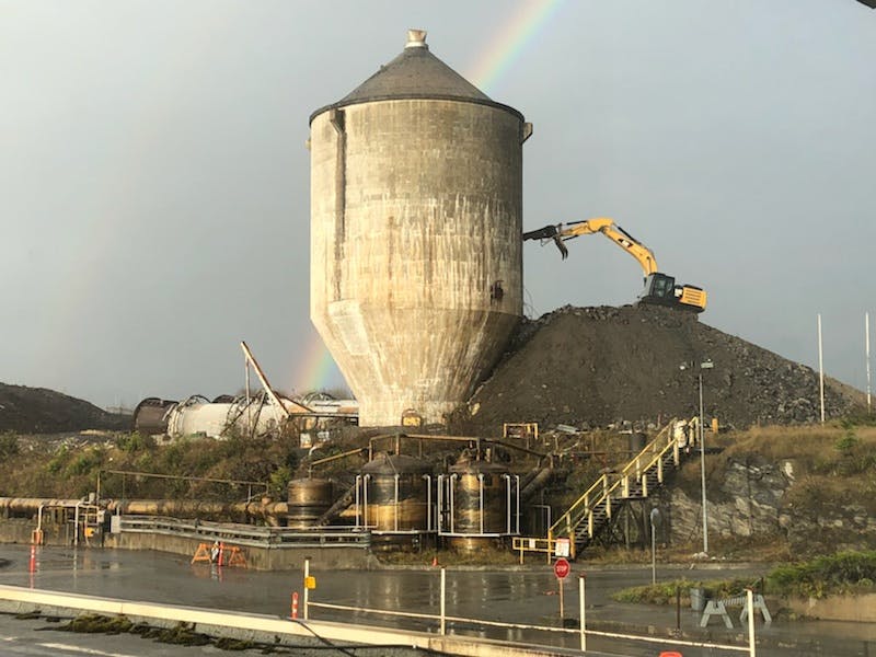 A yellow construction machine on a dirt hill beside a large concrete tower in front of a grey sky with a rainbow.